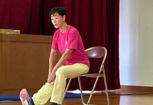 椅子に座り、伸ばした右足の膝を手で押さえる体操をする講師の写真