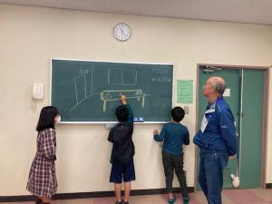 ３人の子供が答えを黒板に描いている様子と見守る先生の写真