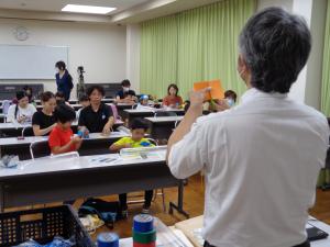 画用紙の切り方を説明させる講師先生と熱心に聞く参加者の様子