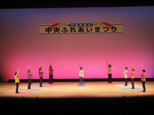 立位で左手を右肩に、右手を真上に伸ばすヨガを披露されるグループの皆さん