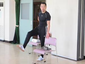 講師が椅子の右側に立ち背もたれに左手を置いて右足をあげて体操の説明をする様子