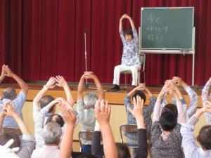 舞台の上で、両手を組んで上に上げる運動を指導する講師と、講師と合わせて両手を上げている受講生の皆さん
