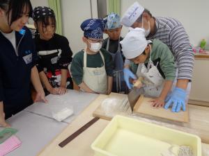スタッフの手助けをうけながら包丁で麺を切る男の子とそれを心配そうに見守る児童たち