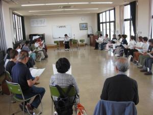歌を指導している講師と、輪になって座って歌っている受講生の様子