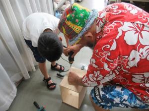 電動ドライバーを使って２枚の板をねじで留めていく作業の指導を受ける小学生と講師