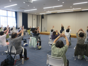 椅子に座って頭の上で両手を重ねる。肩と肩甲骨を滑らかに動かすトレーニングをする受講生