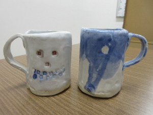 白地に淡い青色のマグカップ２個。子ども作品はオバケの顔を釉薬の色を変えて強調しています。