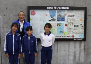 掲示板の横に立つ小谷会長と３名の九会小学生